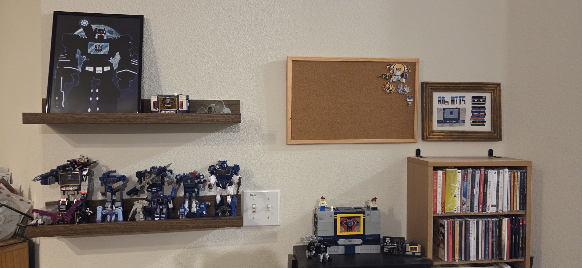 Soundwave toys and art displayed on a wall Picture of a wall that has a couple shelves that have various Soundwave toys and a framed print, CD shelving where a LEGO Soundwave sits, a corkboard with some pins, and the framed cross stitch piece.